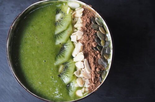 Close up picture of a green smoothie in a coconut bowl topped with slices of kiwi, flaked almonds, cacoa powder and pumpkin seeds