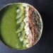 Close up picture of a green smoothie in a coconut bowl topped with slices of kiwi, flaked almonds, cacoa powder and pumpkin seeds