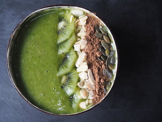 Close up picture of a green smoothie in a coconut bowl topped with slices of kiwi, flaked almonds, cacoa powder and pumpkin seeds